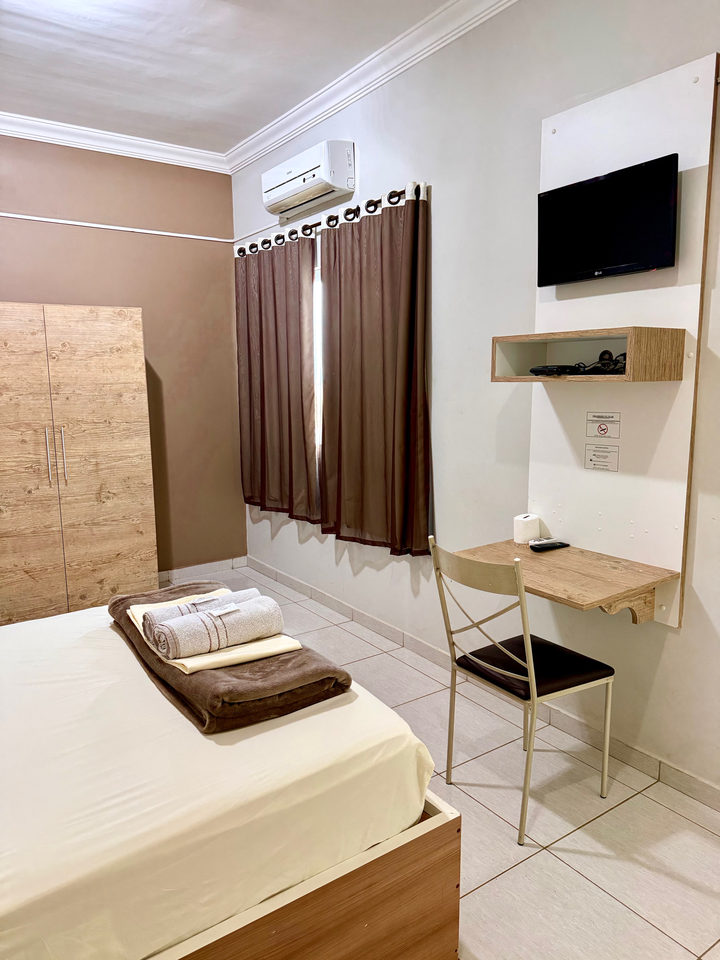 Cama de casal com televisão na parede, escrivaninha e guarda-roupa