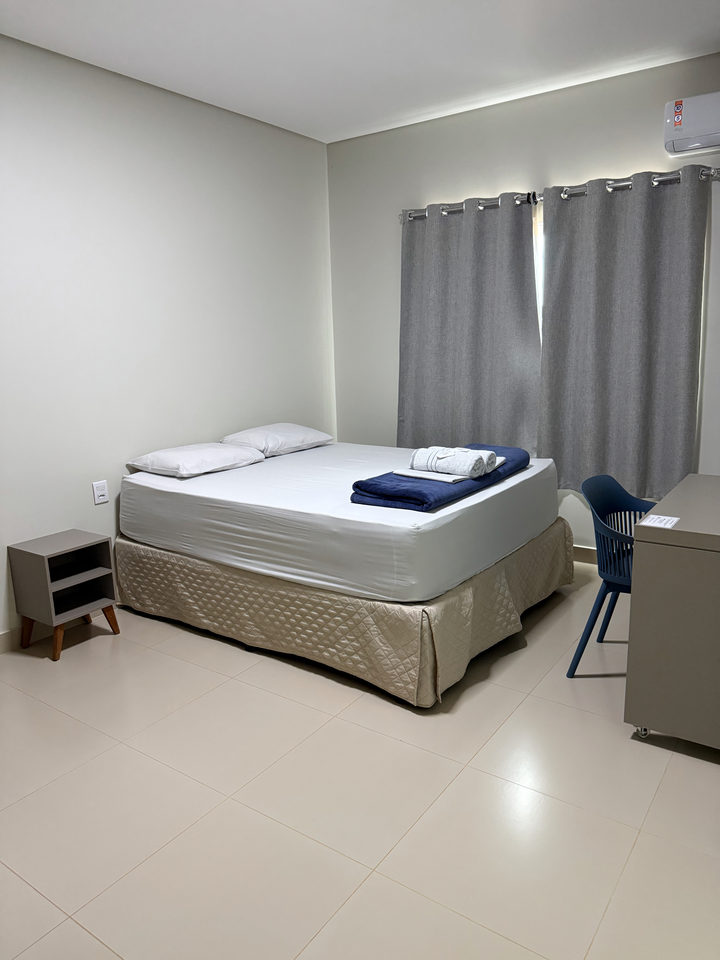 Cama queen com cortina cinza, criado-mudo, escrivaninha e cadeira azul