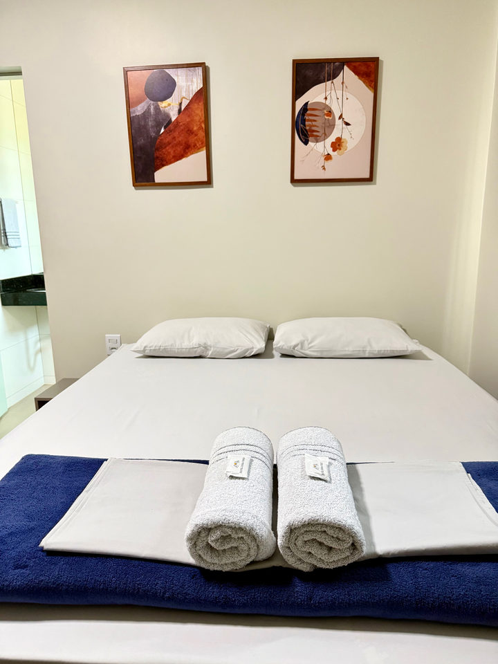 Cama queen com enxoval azul, toalhas enroladas e dois quadros